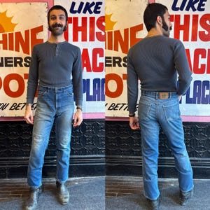 May include: A person wearing a gray long-sleeved Henley shirt and blue jeans. The jeans have a faded wash and distressed details. The person is also wearing black boots. The background features a vintage poster.
