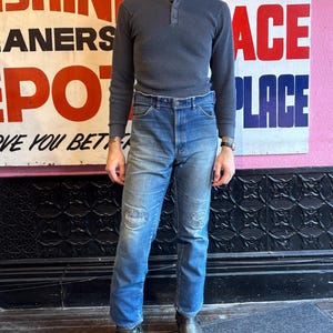 May include: A person wearing a gray waffle-knit Henley shirt and blue jeans with distressed patches. The person is wearing gray leather boots. A vintage sign is in the background.