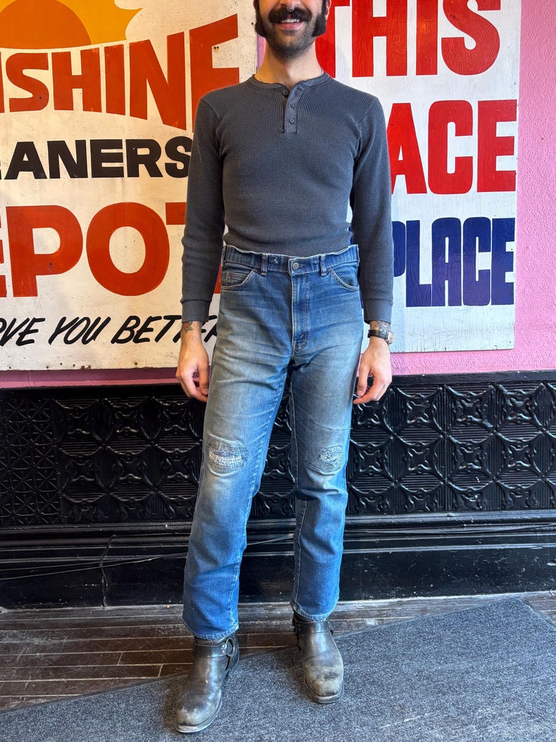 May include: A person wearing a gray waffle-knit Henley shirt and blue jeans with distressed patches. The person is wearing gray leather boots. A vintage sign is in the background.