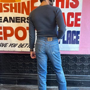 May include: A person wearing a dark gray waffle-knit long-sleeve shirt and light blue jeans. The jeans have a brown leather belt with a logo. A vintage sign is in the background with the words "Sunshine Cleaners Depot".