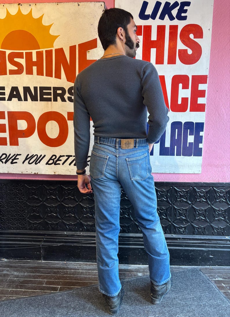 May include: A person wearing a dark gray waffle-knit long-sleeve shirt and light blue jeans. The jeans have a brown leather belt with a logo. A vintage sign is in the background with the words "Sunshine Cleaners Depot".