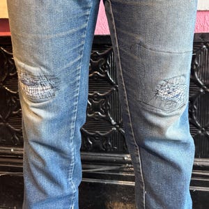 May include: Faded blue denim jeans with distressed patches on the knees. The jeans have a classic five-pocket design and a straight leg cut. The patches are stitched with white thread, adding a unique detail to the garment.