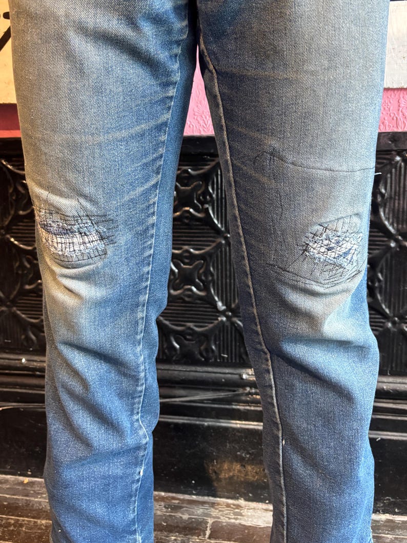May include: Faded blue denim jeans with distressed patches on the knees. The jeans have a classic five-pocket design and a straight leg cut. The patches are stitched with white thread, adding a unique detail to the garment.
