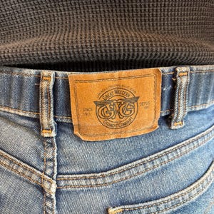 May include: Close-up of blue denim jeans with a brown leather patch. The patch reads "Great Western Garment Company" with text "Canada's Original Blue Jeans" and "Since 1911". The jeans have visible stitching and back pockets.
