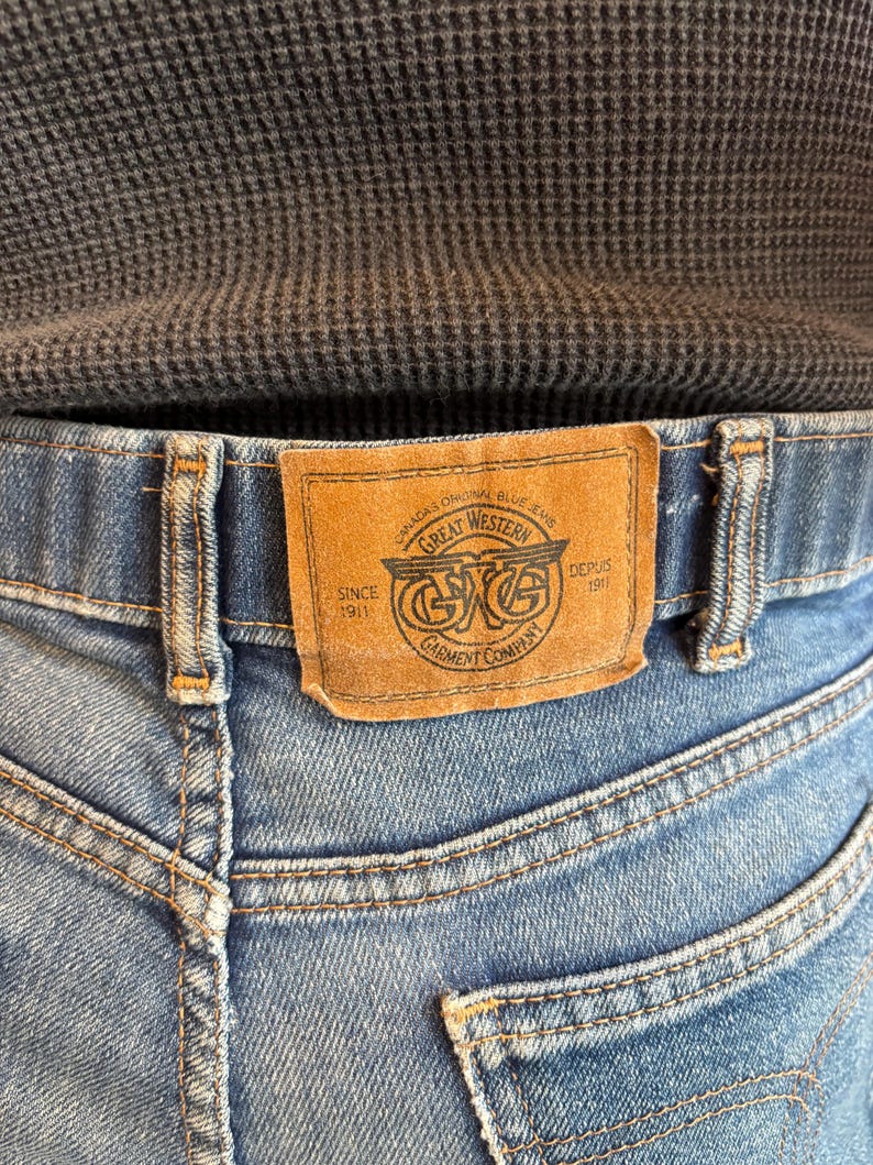May include: Close-up of blue denim jeans with a brown leather patch. The patch reads "Great Western Garment Company" with text "Canada's Original Blue Jeans" and "Since 1911". The jeans have visible stitching and back pockets.