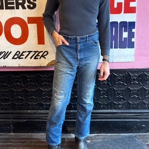 May include: A dark gray waffle-knit Henley shirt with three buttons and long sleeves. The shirt is paired with blue denim jeans with distressed patches on the knees. The outfit is completed with black leather boots.