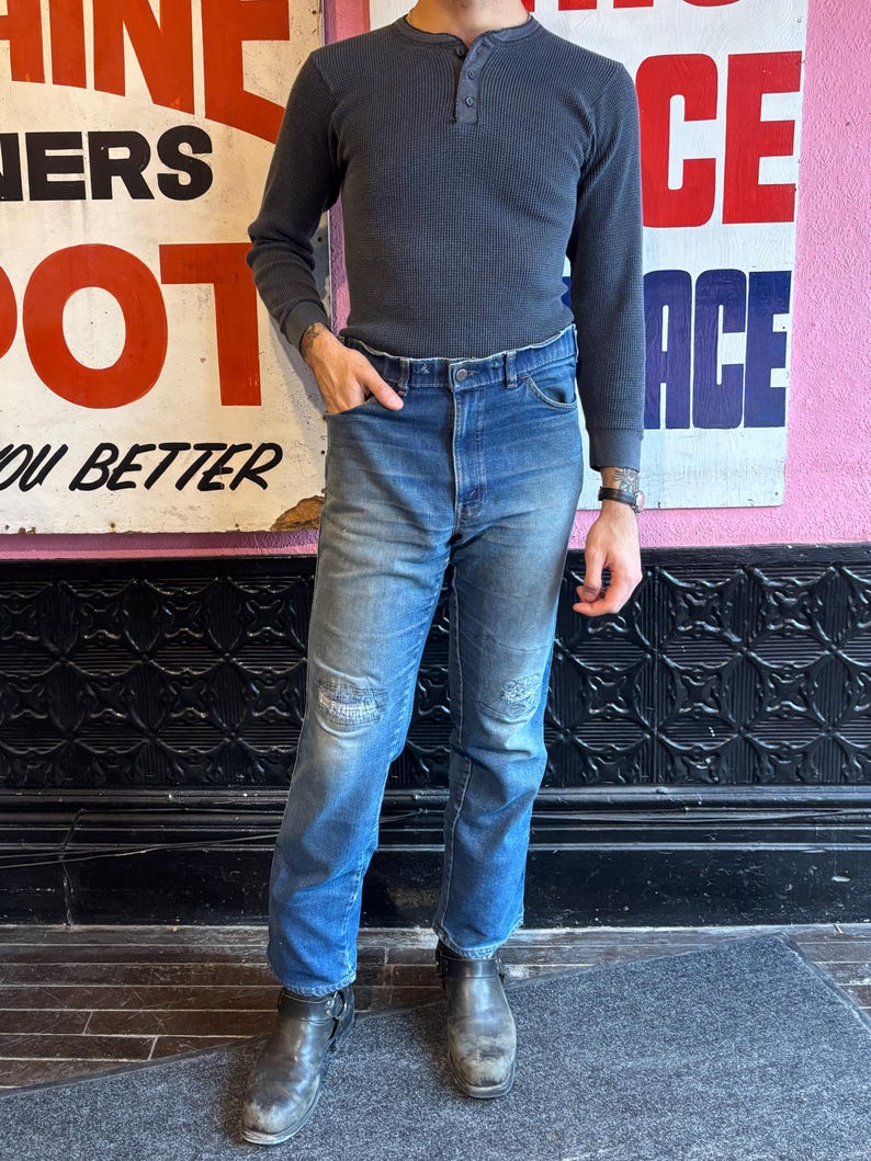 May include: A dark gray waffle-knit Henley shirt with three buttons and long sleeves. The shirt is paired with blue denim jeans with distressed patches on the knees. The outfit is completed with black leather boots.