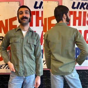 Può includere: Camicia verde oliva con bottoni, due tasche sul petto e maniche lunghe. La camicia ha un colletto classico e una vestibilità rilassata. La camicia è abbinata a jeans azzurri. Stile militare vintage.