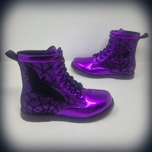 May include: A pair of shiny purple combat boots. The boots feature black laces, a black pull tab, and a black spiderweb design with a spider on one boot and a bat on the other. The boots are a vibrant purple color.