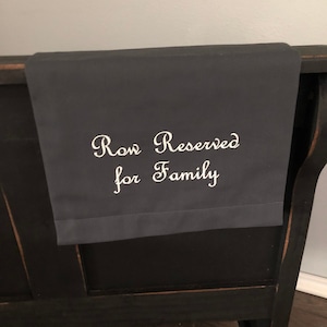 May include: A dark gray fabric banner with the embroidered text "Row Reserved for Family" in white script. The banner is draped over the back of a black wooden bench, suggesting a seating arrangement for family.
