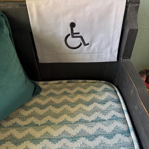 May include: A light gray fabric headrest cover with a black embroidered wheelchair symbol. The cover is placed on a dark wooden chair back. A teal pillow is visible on the left, and the chair seat has a blue and white chevron pattern.