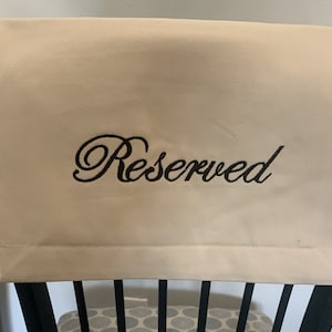May include: A beige chair cover with the word "Reserved" embroidered in black script. The cover is draped over the back of a black chair, indicating a table setting or event seating arrangement.