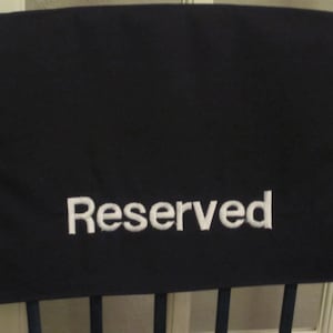 May include: A black chair cover with the word "Reserved" embroidered in white. The cover is rectangular and draped over the back of a wooden chair, indicating a seat is unavailable.