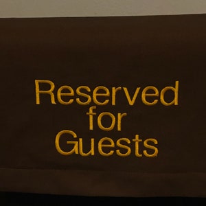 May include: A brown fabric sign with the embroidered text "Reserved for Guests" in gold thread. The sign is rectangular and appears to be hanging on a dark surface, likely a piece of furniture. The text is centered and easy to read.