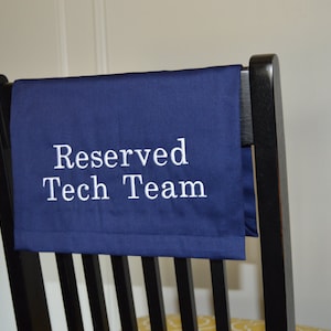 May include: A navy blue chair cover with the text "Reserved Tech Team" embroidered in white. The cover is draped over the back of a black wooden chair. The chair has vertical slats.
