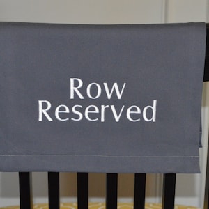 May include: A gray fabric chair cover with the words "Row Reserved" embroidered in white. The cover is draped over the back of a black wooden chair. The background is a light-colored wall.