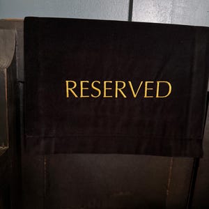 May include: Black velvet chair cover with the word "RESERVED" embroidered in gold thread. The cover is draped over the back of a dark wooden chair, indicating a seat is unavailable.