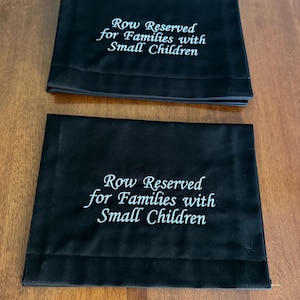 May include: Two black fabric signs with white embroidered text that reads "Row Reserved for Families with Small Children". The signs are rectangular and folded.