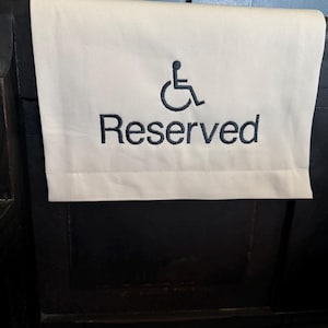 May include: Cream-colored fabric sign with the word "Reserved" embroidered in dark blue, along with the wheelchair symbol. The sign is draped over a dark wooden bench, indicating a reserved seating area.