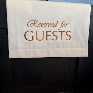 May include: A white rectangular cloth with the words "Reserved for GUESTS" embroidered in a gold-brown color. The cloth is folded over a dark surface, likely a table or chest.