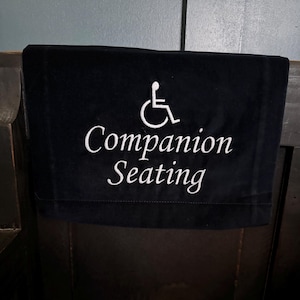 May include: A black fabric sign with white embroidered text and a wheelchair symbol. The sign reads "Companion Seating" and is draped over a dark wooden surface.