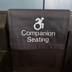 May include: A black fabric sign with a white wheelchair accessibility symbol and the text "Companion Seating".