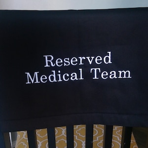 May include: Black fabric with white embroidered text that reads "Reserved Medical Team." The fabric is draped over the back of a chair with a yellow and white patterned cushion visible below.