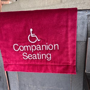 May include: A burgundy velvet seat cover with white embroidered text and a wheelchair symbol. The text reads "Companion Seating". The cover is draped over the back of a dark wooden chair.