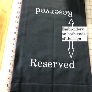 May include: A dark gray fabric sign with the word "Reserved" embroidered in white on both ends. The sign is rectangular and appears to be made of a durable material, suitable for repeated use.