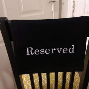 May include: A black chair with a black fabric draped over the back, displaying the word "Reserved" in white. The seat cushion has a yellow and white pattern. The chair is in front of a white door.