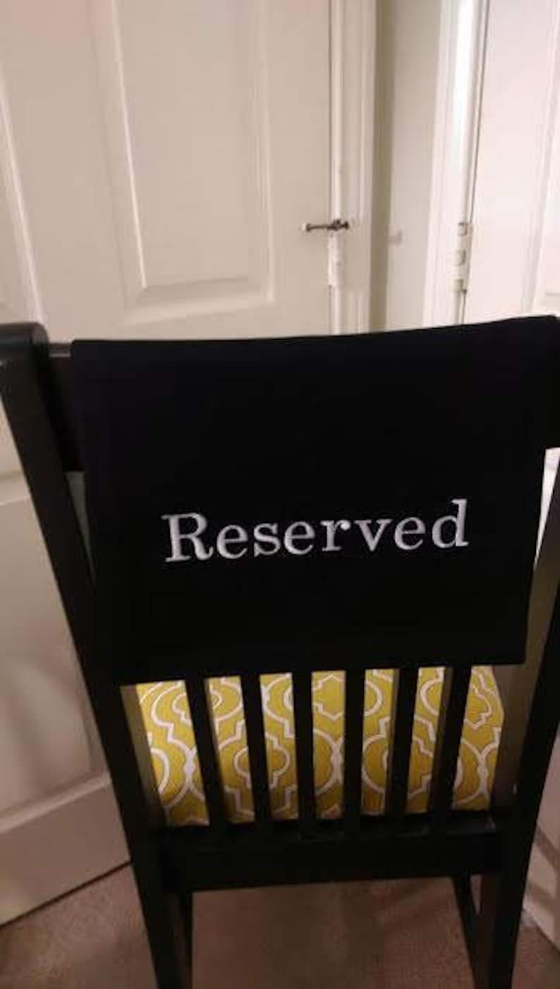 May include: A black chair with a black fabric draped over the back, displaying the word "Reserved" in white. The seat cushion has a yellow and white pattern. The chair is in front of a white door.