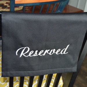 May include: Black chair cover with the word "Reserved" embroidered in white script. The cover is draped over the back of a black chair. The chair is in a room with a yellow and white patterned chair cushion.