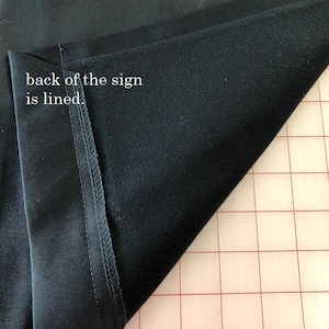 May include: Close-up of a black fabric with a lined interior. The text "back of the sign is lined" is visible. The fabric is folded, revealing the interior lining and the stitching along the edge.