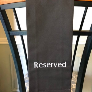 May include: A dark brown fabric towel with the word "Reserved" embroidered in white. The towel is draped over the back of a black chair with a wooden top. The chair has a patterned cushion.