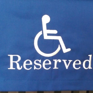 May include: A blue sign with white text and a wheelchair symbol. The word "Reserved" is embroidered below the symbol. The sign is likely used to indicate a reserved parking space or seating area for individuals with disabilities.