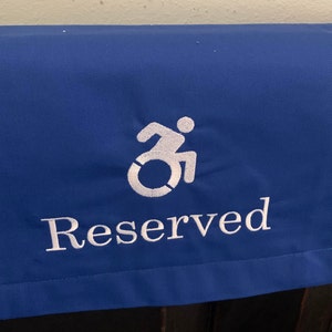 May include: A royal blue fabric cover with the word "Reserved" embroidered in white, along with the wheelchair symbol. The cover is designed to be placed over a seat back.