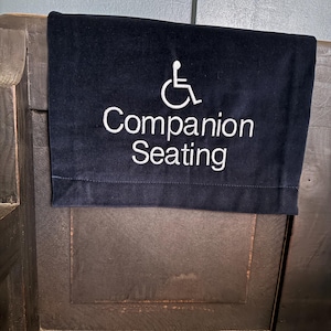 May include: A dark blue fabric sign with white embroidered text and a wheelchair symbol. The sign reads "Companion Seating" and is displayed on a wooden surface.