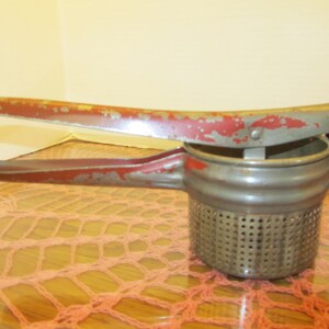 May include: Vintage potato ricer with a red and silver finish. The handle and frame show signs of wear, with chipped red paint. The ricer has a perforated bowl for mashing potatoes and other vegetables. A classic kitchen tool.