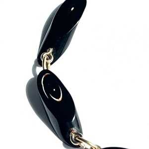 May include: A close-up of a black necklace with oval-shaped beads. The beads are connected by gold-colored chain links. Each bead has a small, light-colored design. The necklace is set against a white background.