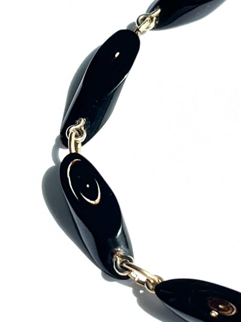 May include: A close-up of a black necklace with oval-shaped beads. The beads are connected by gold-colored chain links. Each bead has a small, light-colored design. The necklace is set against a white background.