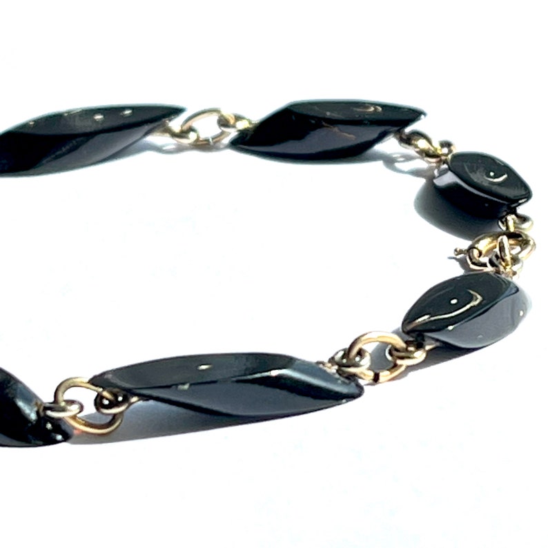 May include: A vintage bracelet featuring black, teardrop-shaped beads linked by gold-tone metal chain. The beads have a glossy finish, and the chain has a delicate design. The bracelet is set against a white background.