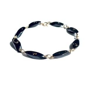 May include: A black beaded bracelet with oval-shaped beads. The beads are linked together with small gold-colored chain links. The bracelet is set against a white background.
