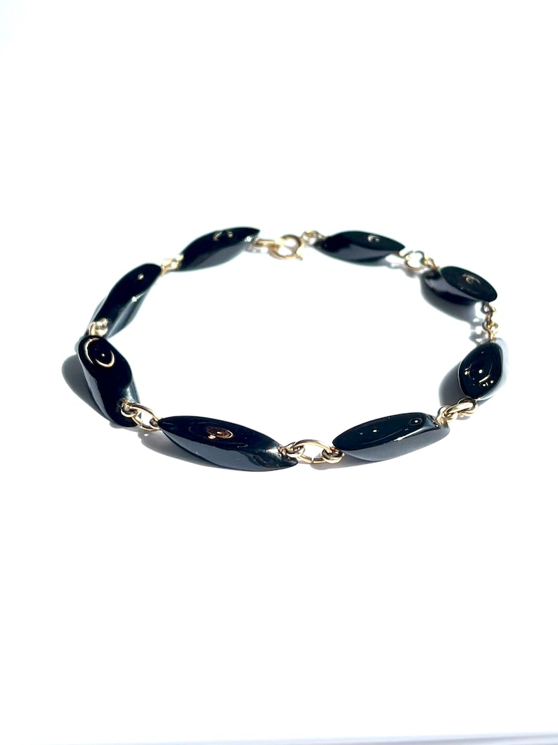 May include: A black beaded bracelet with oval-shaped beads. The beads are linked together with small gold-colored chain links. The bracelet is set against a white background.