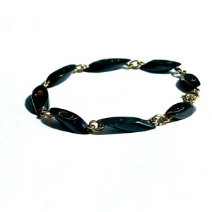 May include: A vintage bracelet featuring elongated, black, oval-shaped beads linked by small gold-colored metal chains. The bracelet is displayed on a white background, highlighting its glossy, dark beads and the contrast with the gold-toned metal.