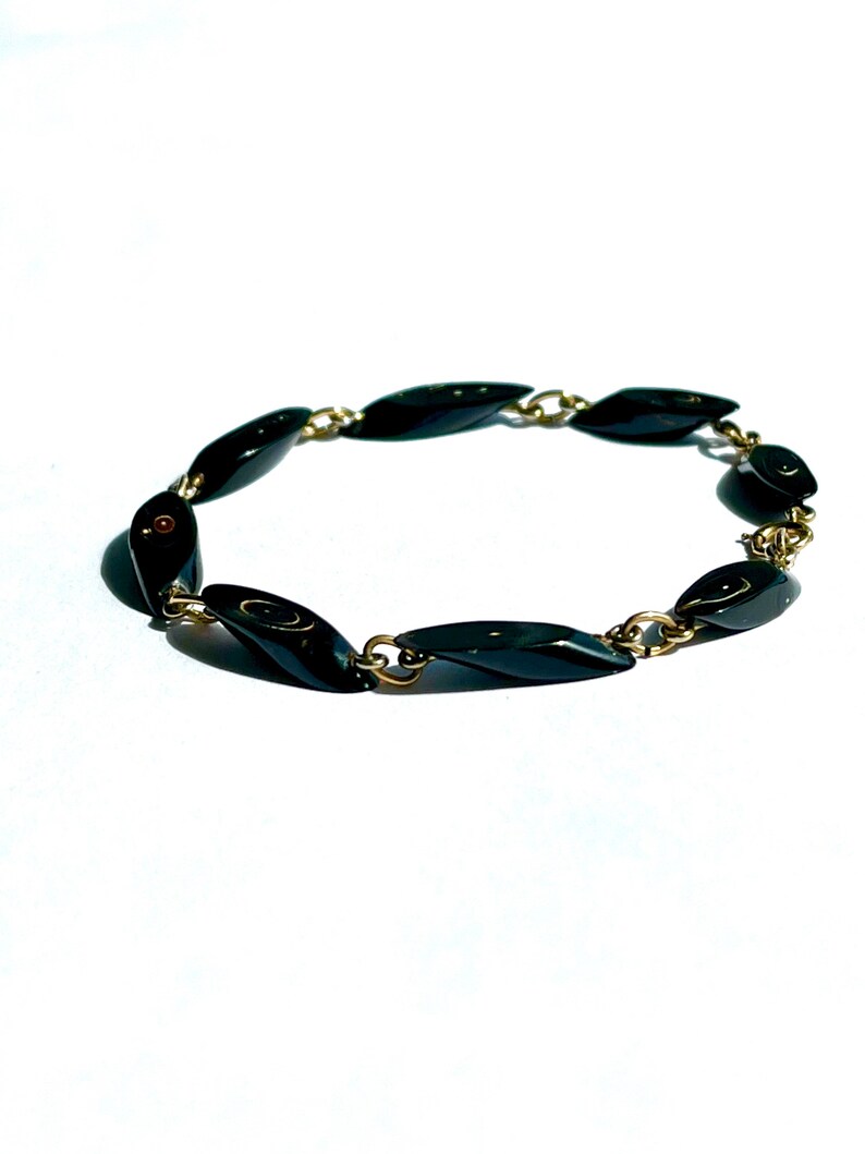 May include: A vintage bracelet featuring elongated, black, oval-shaped beads linked by small gold-colored metal chains. The bracelet is displayed on a white background, highlighting its glossy, dark beads and the contrast with the gold-toned metal.