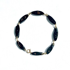May include: A gold-toned bracelet featuring oval-shaped black beads. The beads are linked together with small gold-colored chains and a clasp. The bracelet is displayed on a white background.