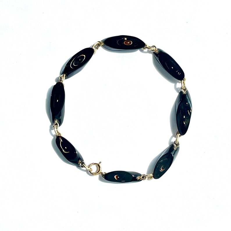 May include: A gold-toned bracelet featuring oval-shaped black beads. The beads are linked together with small gold-colored chains and a clasp. The bracelet is displayed on a white background.