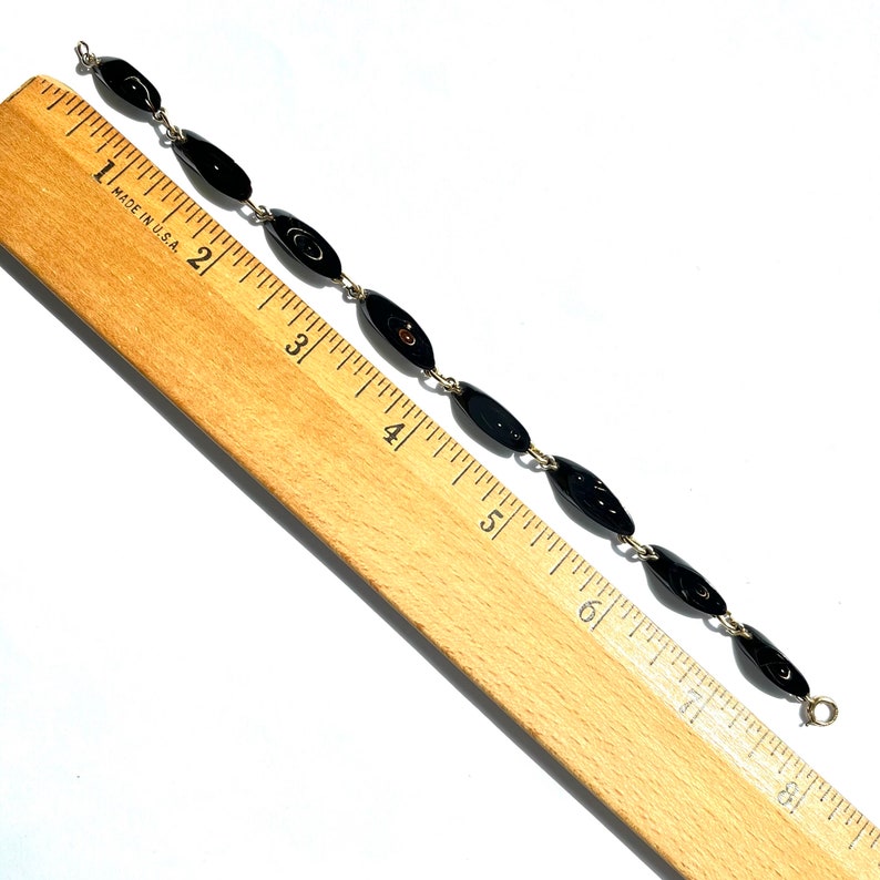May include: A vintage bracelet featuring oval black beads linked by silver-tone metal. The beads are approximately 0.75 inches long. The bracelet is displayed on a wooden ruler, showing measurements in inches, against a white background.