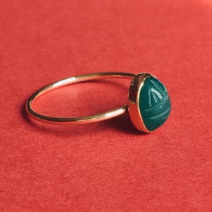 May include: A gold ring featuring a smooth, rounded band and a single, oval-shaped green stone setting. The green stone is carved with a subtle, incised geometric pattern on its surface, held securely within a polished gold bezel setting.