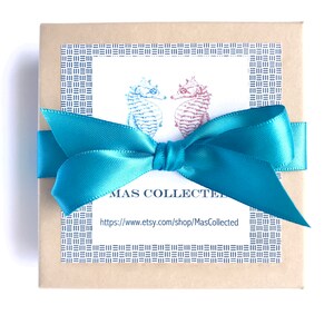 May include: A square, tan gift box tied with a bright teal satin ribbon. The box has a white label with two seahorse illustrations and the text "MAS COLLECTED" and a website address. The label is framed with a blue pattern.